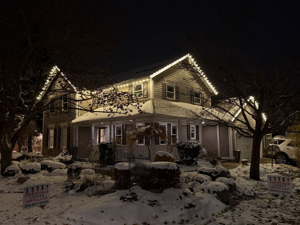 Two-storey home with warm white lights along every roofline, in fresh snow, with Gecko lawn signs